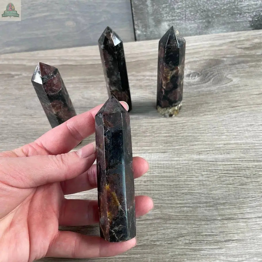 A hand holds a Keystone Crystals Coffee Bean Jasper Gemstone Obelisk (4–5 oz), polished with brown and reddish tones, while three similar bulk crystal towers stand upright on a light wooden surface in the background.