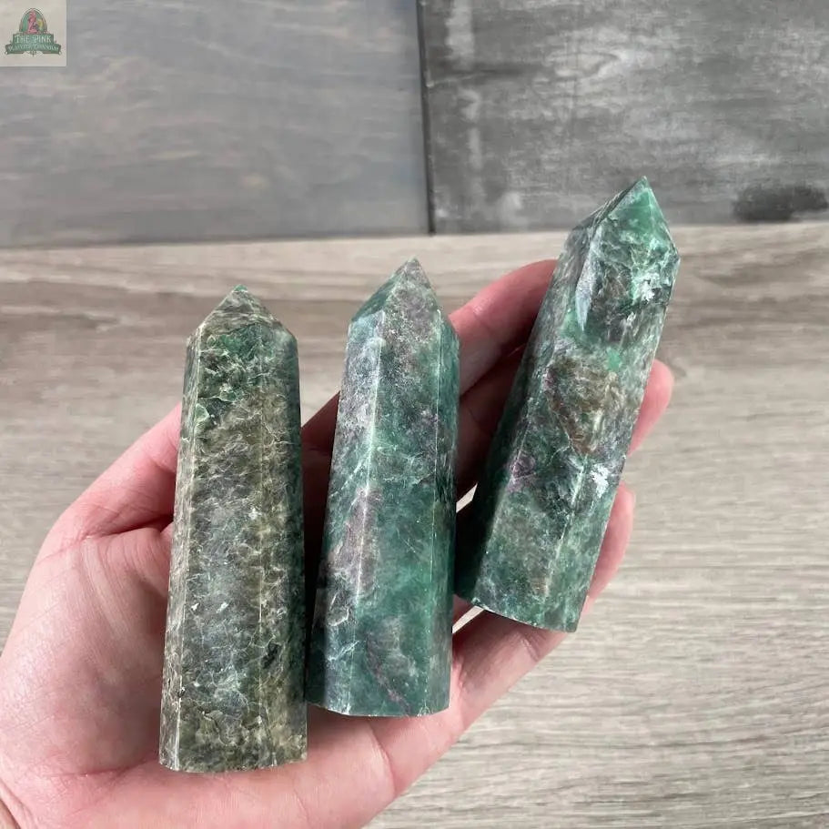 A hand displays three polished green fluorite crystal towers with pointed tips on a wooden surface—ideal for pairing with Keystone Crystals’ Gemstone Obelisks 4–5 oz Bulk Crystal Towers in Coffee Bean Jasper.