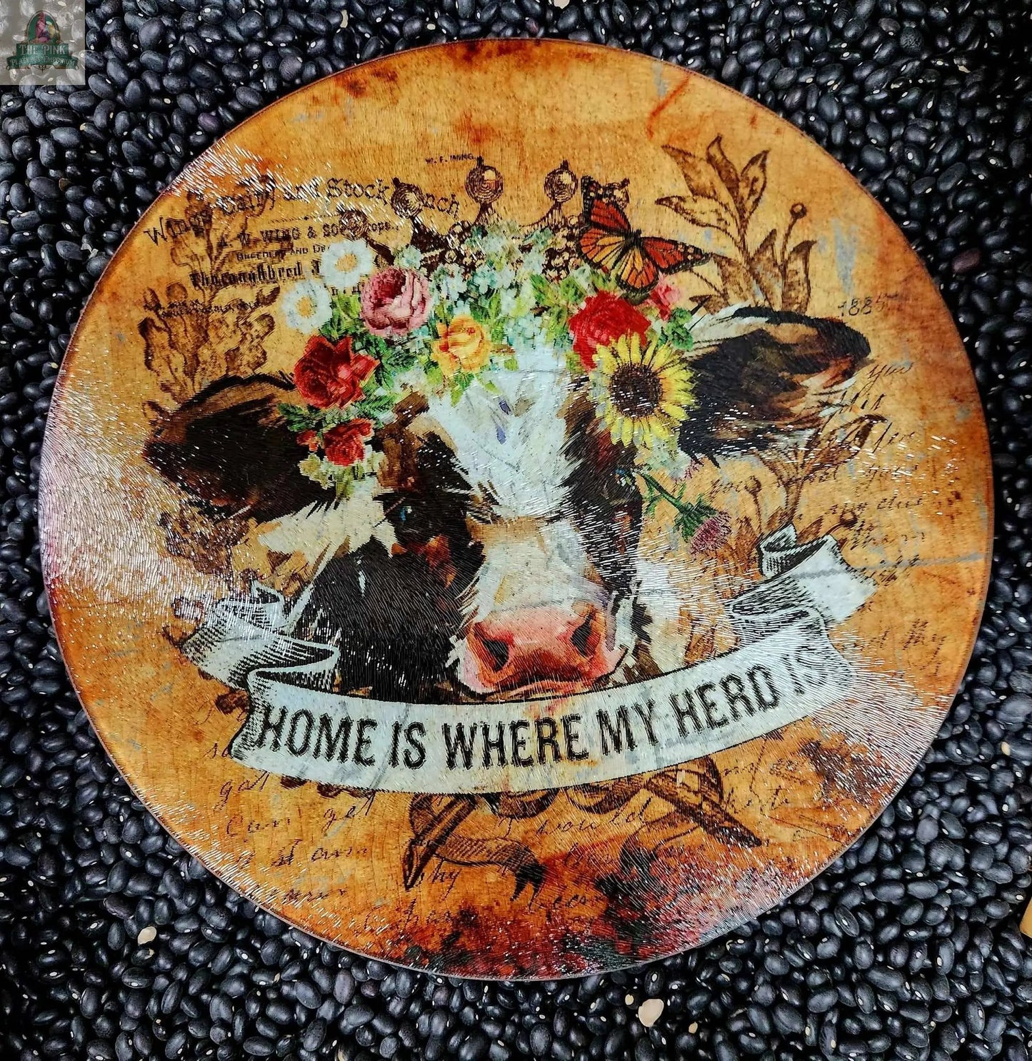 This large 12" glass cutting board, charcuterie, or trivet features a floral cow design with butterflies and foliage, a rustic background, and the phrase "Home is where my herd is." Perfect as a decorative or functional kitchen piece.