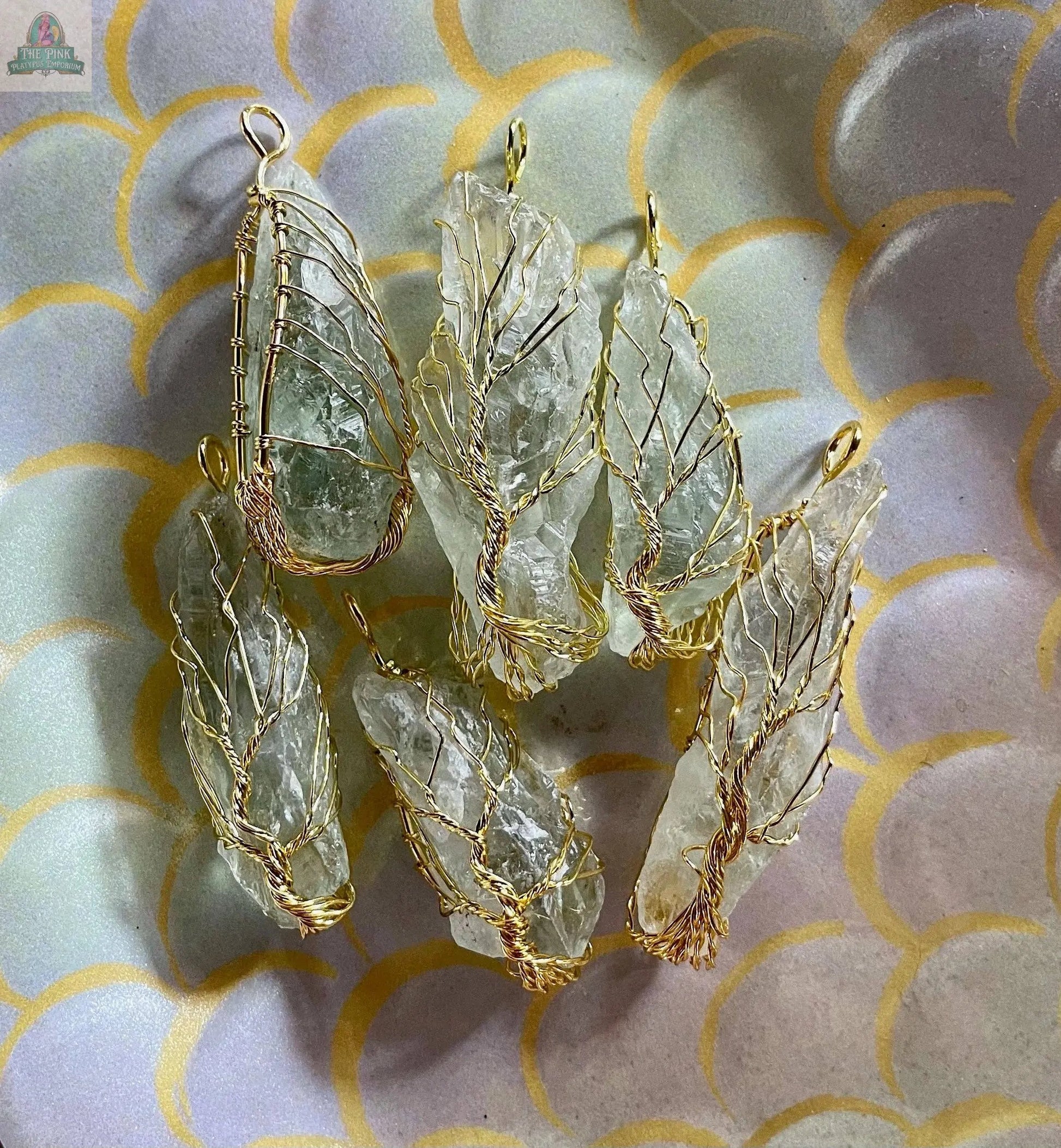 Six rough, translucent crystals encased in gold wire form a cluster for the Golden Wire Wrapped Tree of Life Pendant with Crystals, displayed on a pastel scallop-patterned background. Each pendant features a loop for hanging.