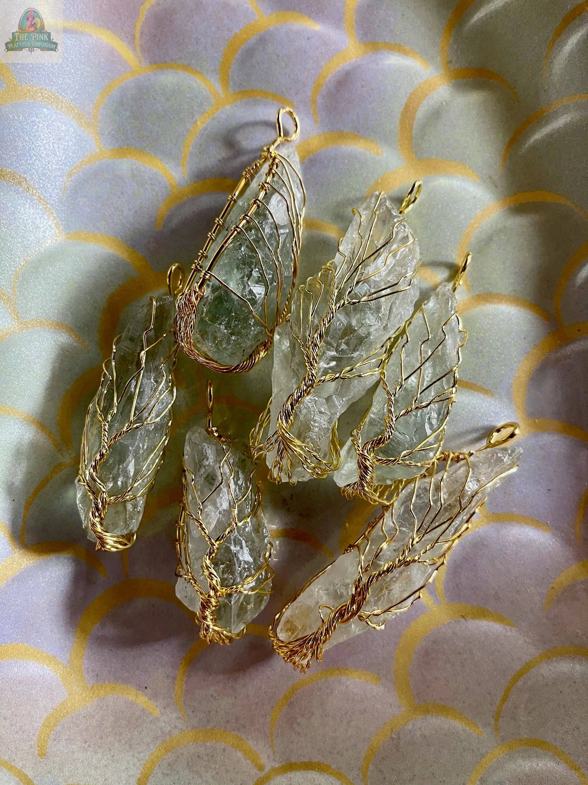 The Golden Wire Wrapped Tree of Life Pendant features rough clear quartz and prasiolite crystals in delicate gold wire. Displayed on a pastel, scalloped-patterned surface with gold accents, it makes a stunning accessory.
