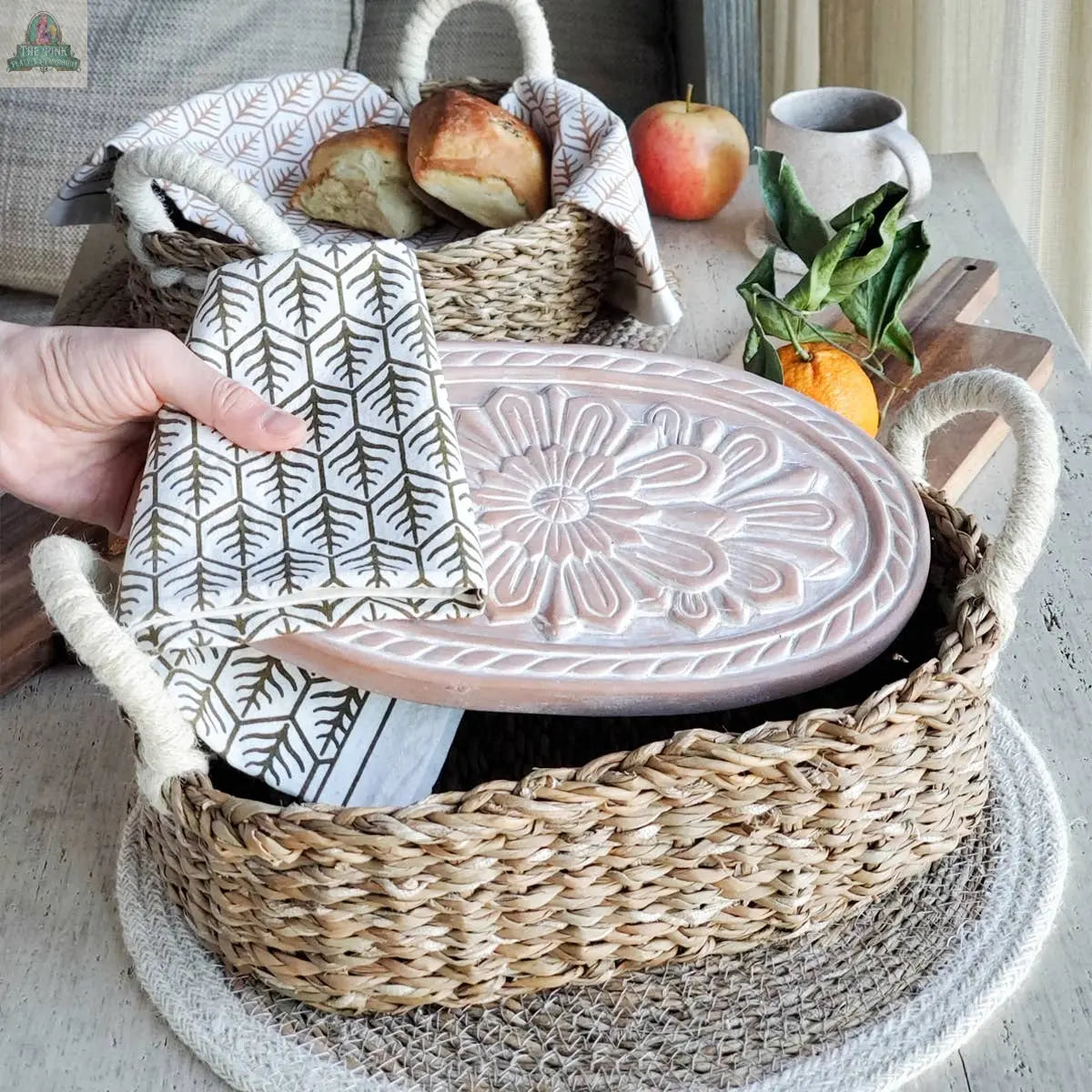 A hand lifts the KORISSA Handmade Bread Warmer & Wicker Basket - Flower from a rustic wooden table, revealing a napkin inside. Nearby are bread, an apple, an orange with leaves, and a cup.