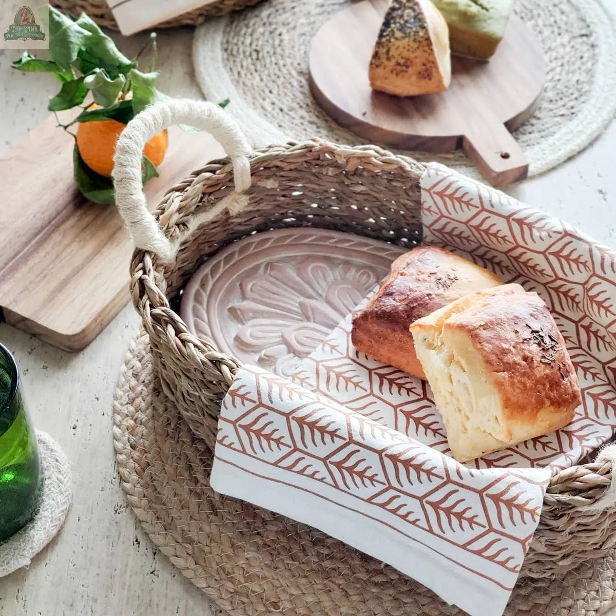 The KORISSA Handmade Bread Warmer & Wicker Basket - Flower holds two breads atop a patterned cloth, resting on a textured placemat, with extra bread and a green drink on the wooden meal table.