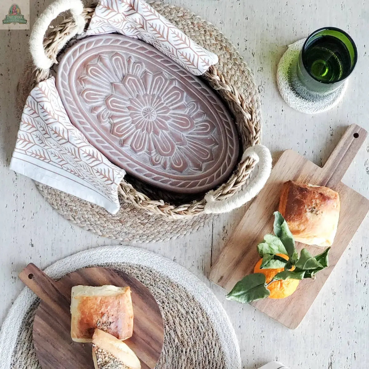 KORISSA Handmade Bread Warmer & Wicker Basket - Flower is paired with a patterned ceramic tray, napkin, wooden bread boards, citrus branch, and a green glass on textured placemats atop a light table.