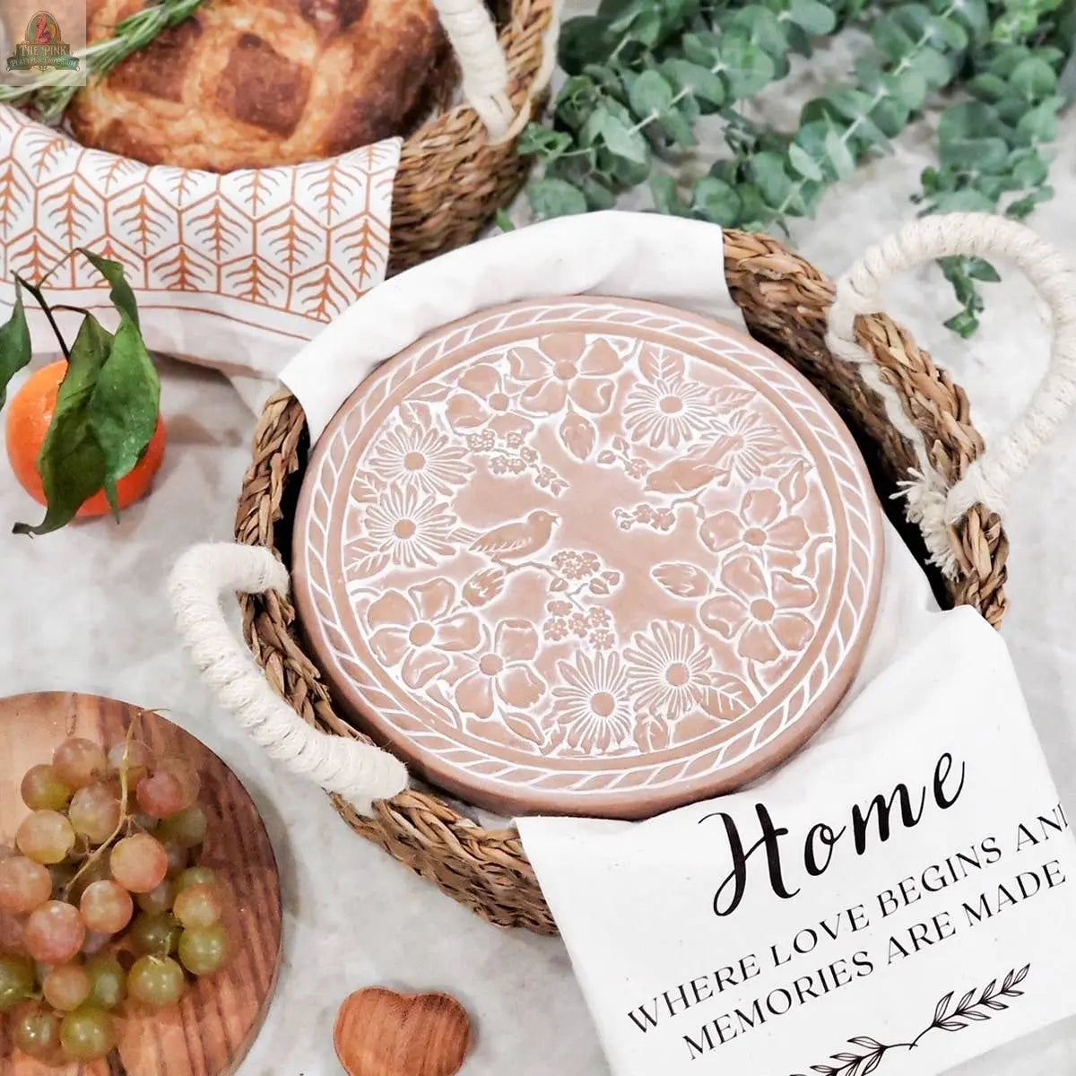 The KORISSA Handmade Bread Warmer & Wicker Basket - Garden Round features a white cloth with the words “Home, where love begins and memories are made” and a terracotta bread warmer with floral designs, perfect for serving fresh bread.
