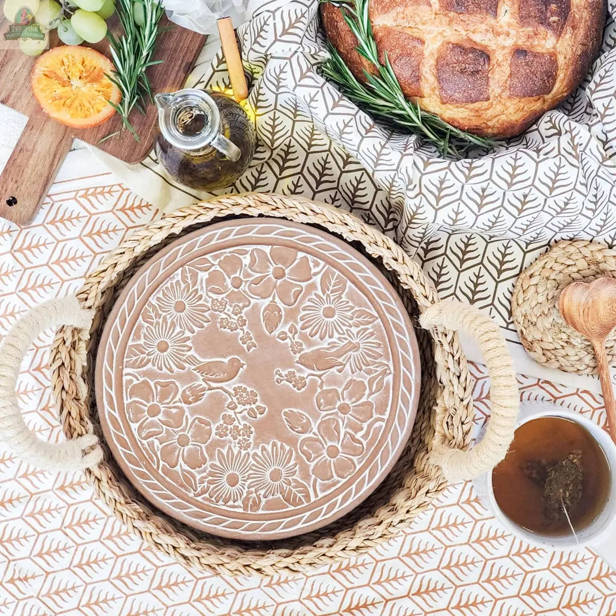The KORISSA Handmade Bread Warmer & Wicker Basket - Garden Round, featuring a floral design, rests on a table alongside bread, an orange half, oil bottle, herbs, and a cup of tea.