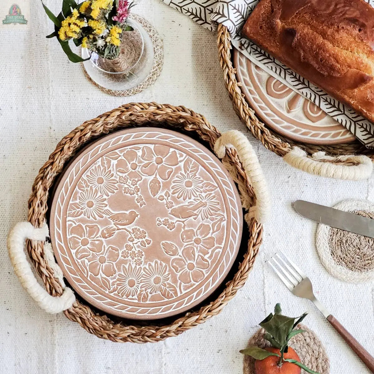The KORISSA Handmade Bread Warmer & Wicker Basket – Garden Round features an engraved terracotta bread warmer in a wicker basket with rope handles, displayed on a beige cloth with bread, cutlery, flowers, and an orange nearby.