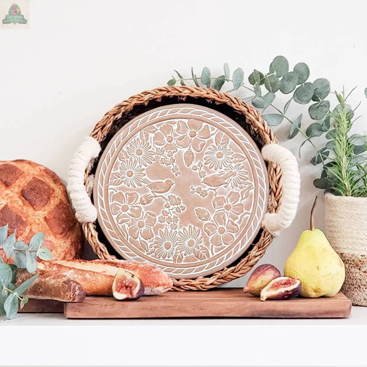 A KORISSA Handmade Bread Warmer & Wicker Basket - Garden Round with white floral patterns displays bread, baguette, figs, a pear, and eucalyptus on a wooden surface. Decorative greenery in a twine-wrapped vase sits behind it.