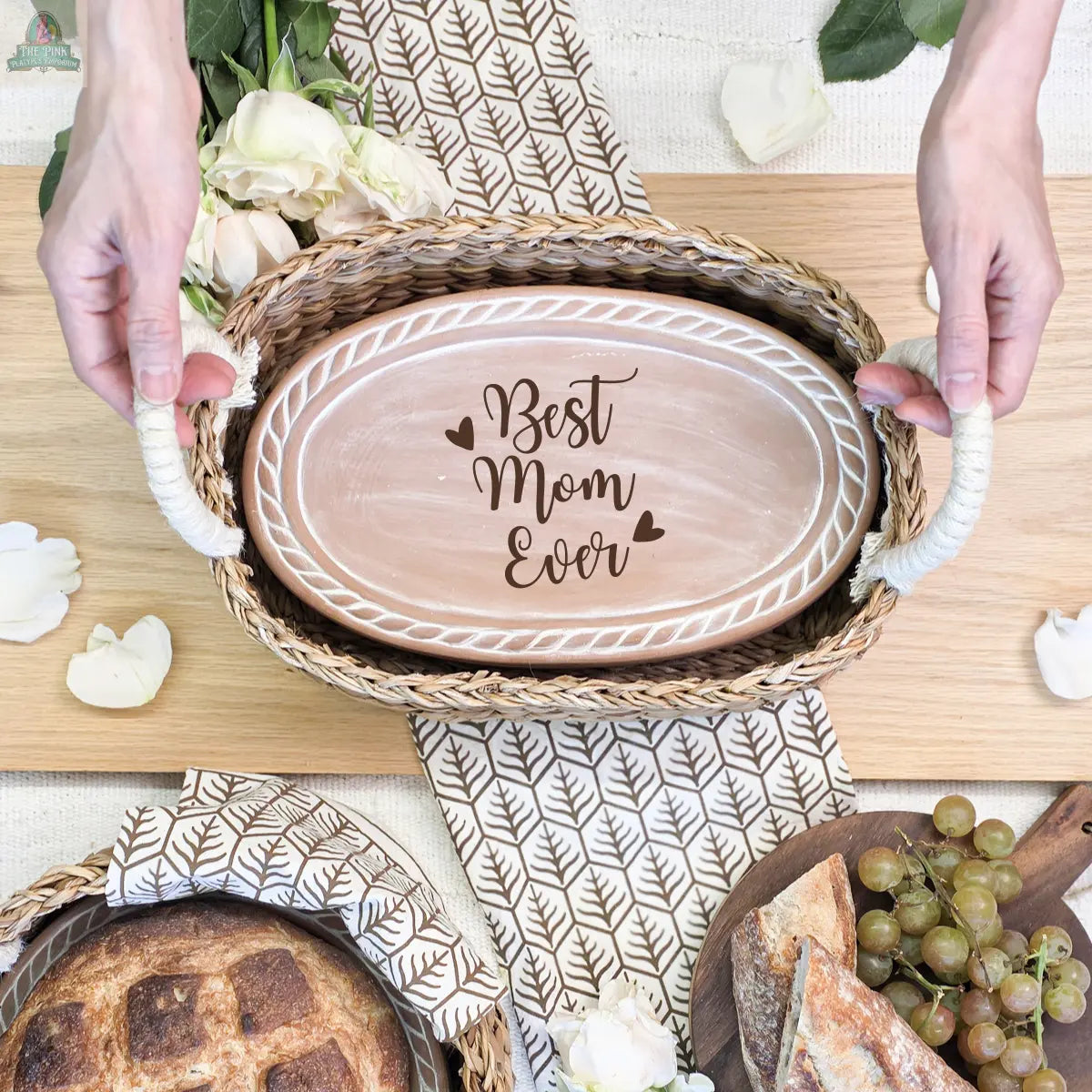 A person holds the Handmade Bread Warmer & Wicker Basket - Mom Oval : Design 3, featuring a decorative "Best Mom Ever" plate, flowers, grapes, napkin, and bread—an ideal bread lover’s gift on a wooden surface.