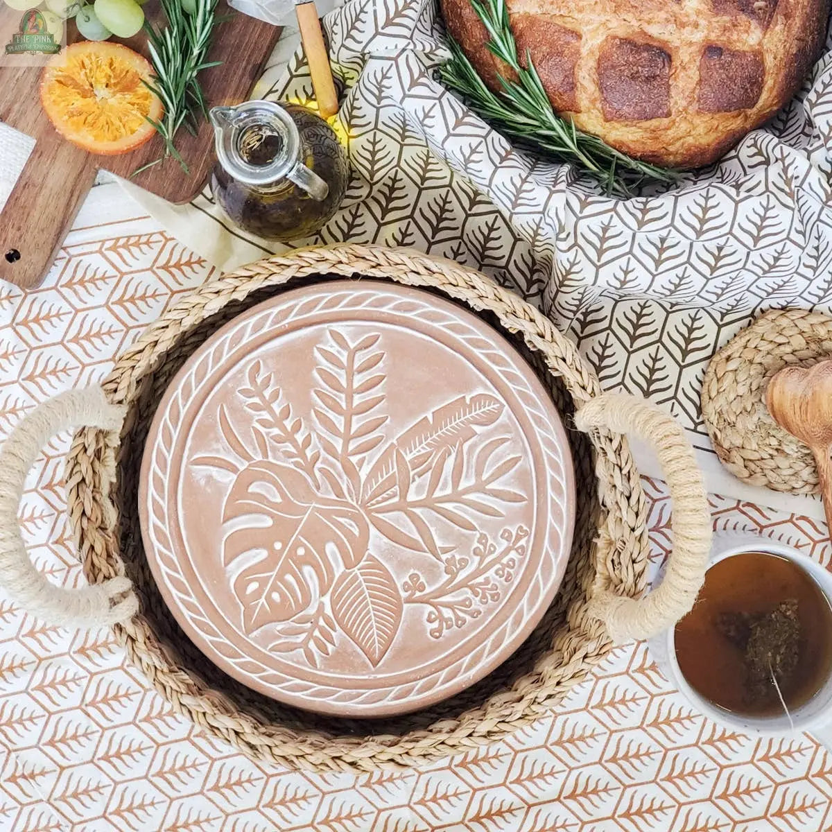 The KORISSA Handmade Bread Warmer & Wicker Basket - Plant Round features a terracotta stone with leaf designs in a wicker basket, perfect alongside fresh bread, tea, oil, fruit slices, and honey on your table.