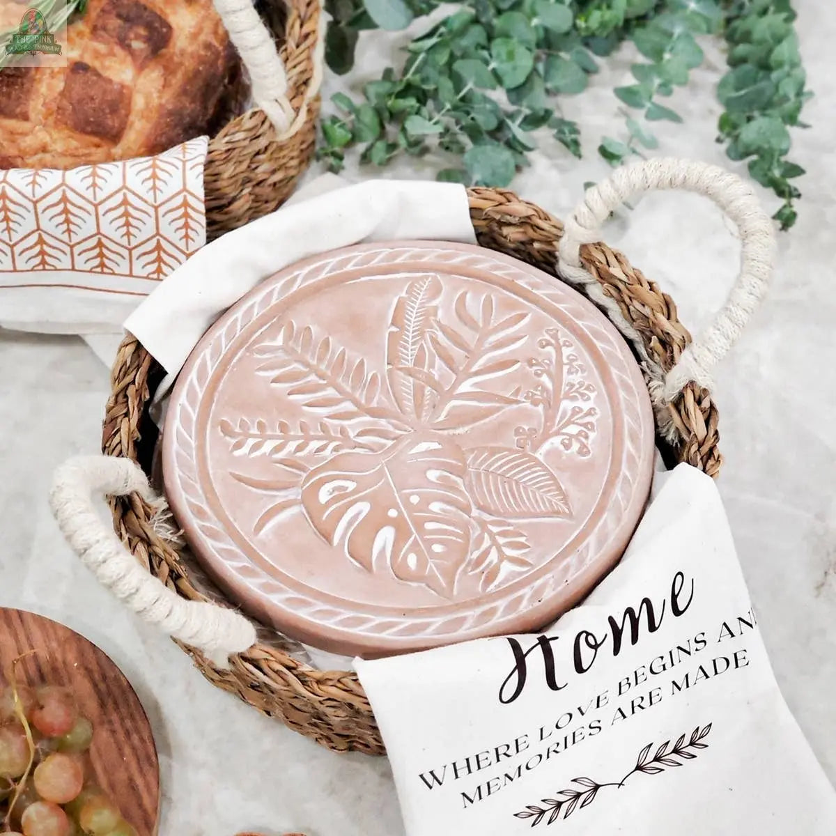 The KORISSA Handmade Bread Warmer & Wicker Basket - Plant Round features a pink ceramic plate with leaf designs, a white cloth reading "Home where love begins and memories are made," plus greenery and bread on a marble surface.