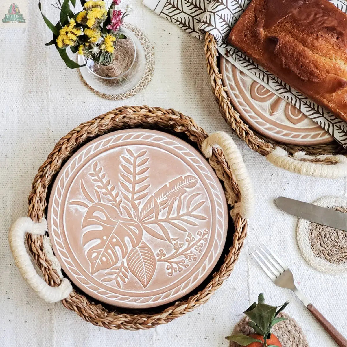 A KORISSA Handmade Bread Warmer & Wicker Basket – Plant Round holds a botanical ceramic plate on a textured cloth, with cutlery, a small flower vase, and sliced fruit beside a loaf on a patterned plate nearby.
