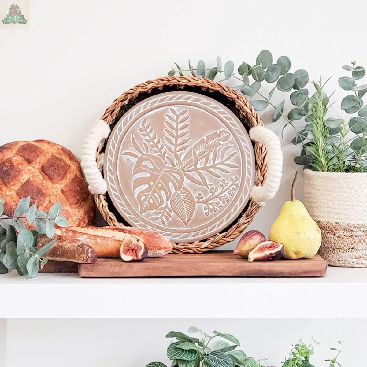 KORISSA’s Handmade Bread Warmer & Wicker Basket - Plant Round sits on a white shelf beside figs, a pear, a baguette, and lush potted plants, creating a fresh, rustic display with a touch of natural charm.