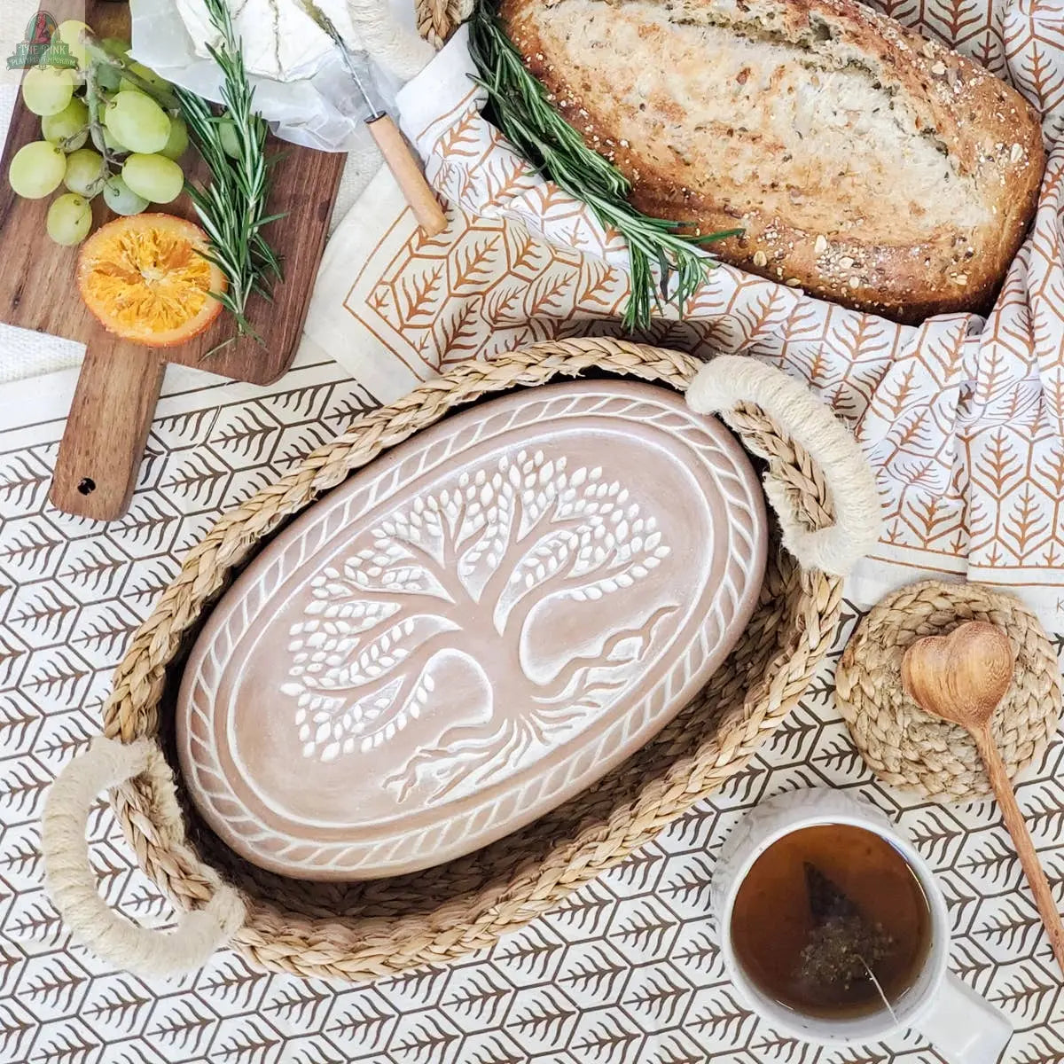 The KORISSA Handmade Bread Warmer & Wicker Basket - Tree of Life Oval keeps bread warm in a ceramic tray featuring a raised tree design, perfect for serving with cheese, fruit, and tea on your favorite patterned tablecloth.