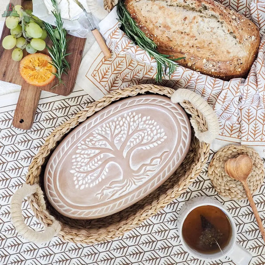 The KORISSA Handmade Bread Warmer & Wicker Basket - Tree of Life Oval keeps bread warm in a ceramic tray featuring a raised tree design, perfect for serving with cheese, fruit, and tea on your favorite patterned tablecloth.