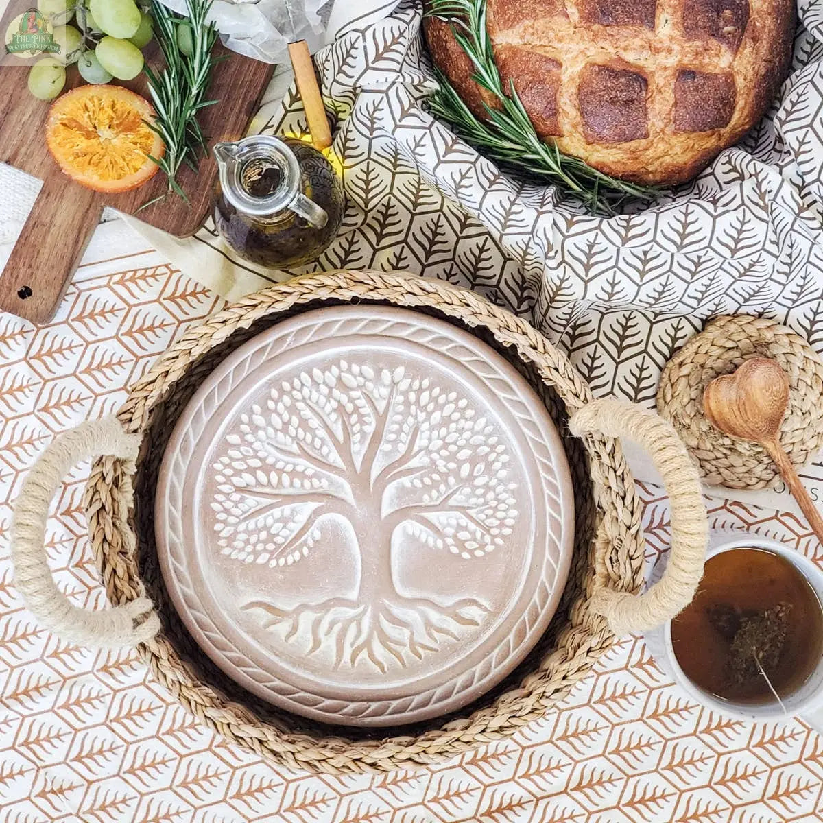 The KORISSA Handmade Bread Warmer & Wicker Basket - Tree of Life Round displays a round loaf with a tree design, surrounded by bread, grapes, sliced orange, oil jar, rosemary, honey dipper, and tea on a patterned cloth.