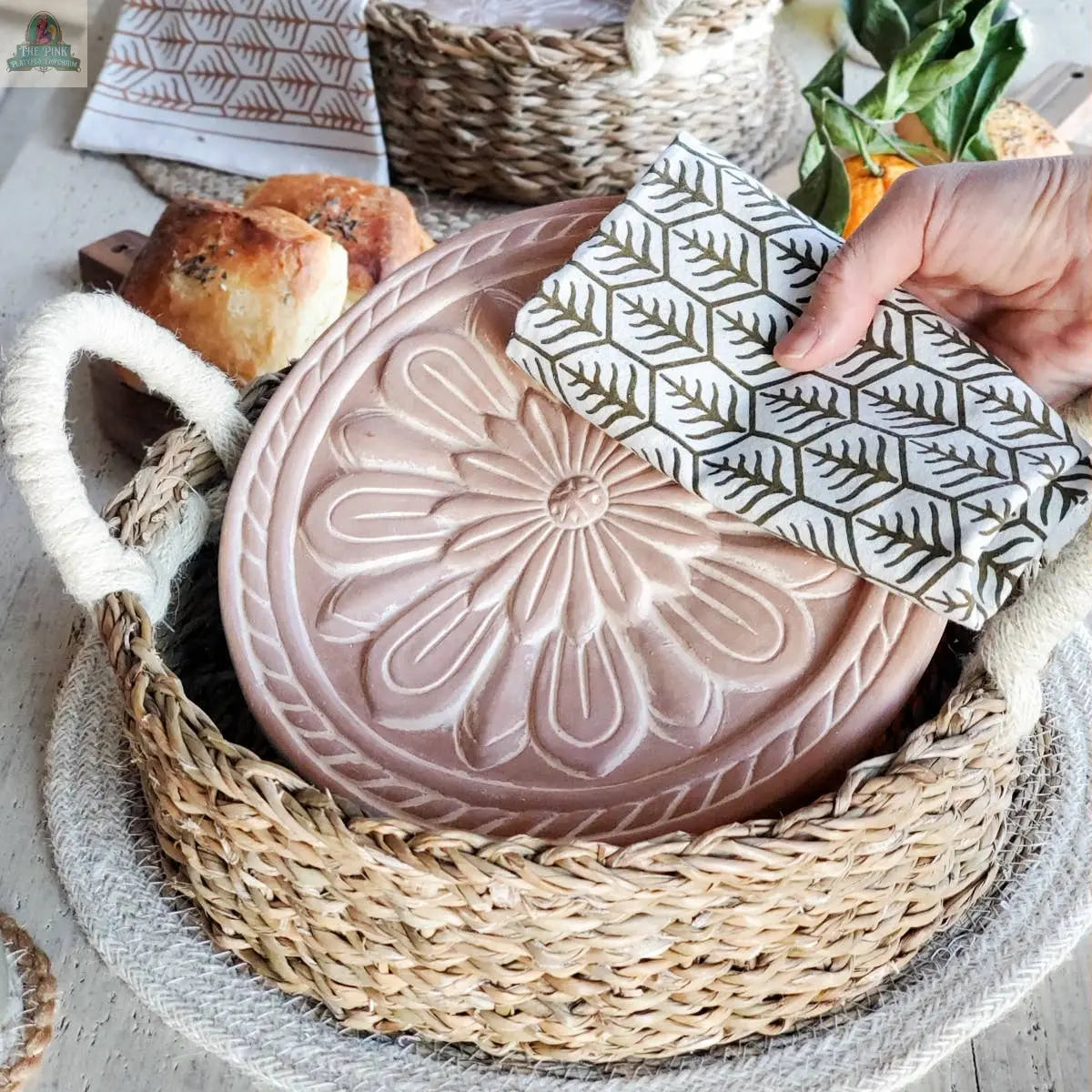A hand drapes a patterned cloth over the KORISSA Handmade Bread Warmer & Wicker Basket - Vintage Flower. Loaves of bread, a pineapple, and another wicker basket sit nearby on a light table.