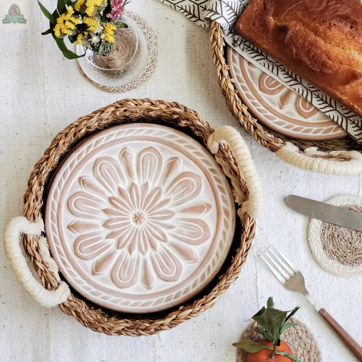 A KORISSA Handmade Bread Warmer & Wicker Basket - Vintage Flower sits beside a round ceramic plate with floral embossing, along with a loaf of bread, utensils, a small flower bouquet, and an orange.