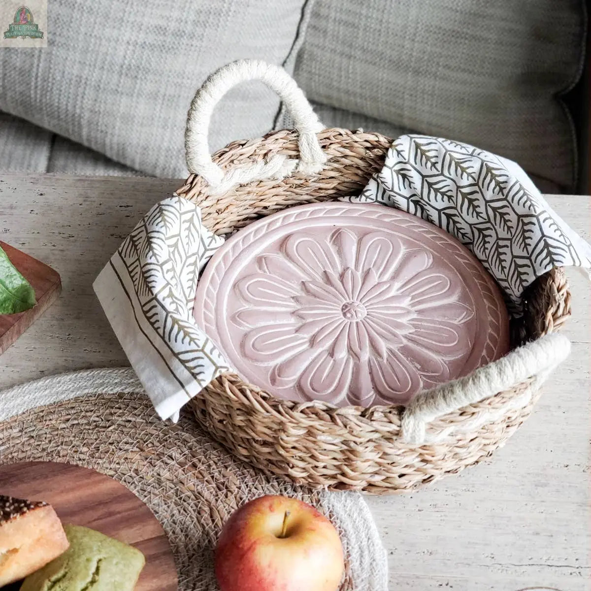 The KORISSA Handmade Bread Warmer & Wicker Basket - Vintage Flower features a round wicker basket with rope handles, a pink floral plate, a white leaf-design cloth, and a terracotta warming stone, displayed on a light wooden table.