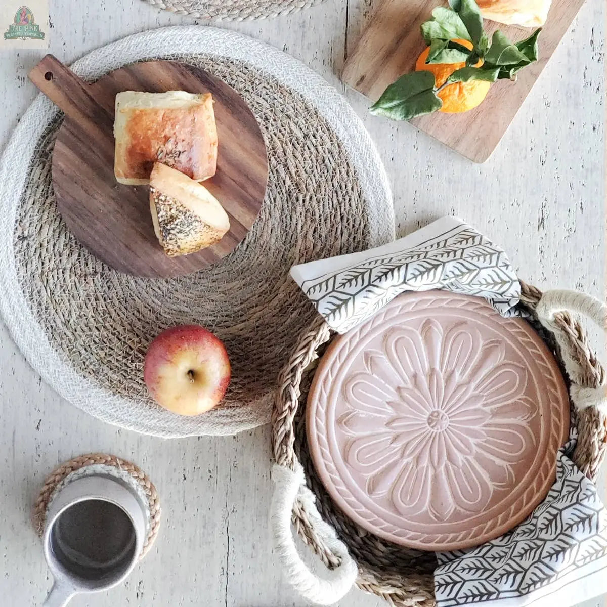 A rustic table features bread on a wooden board, an apple, a mug, and the KORISSA Handmade Bread Warmer & Wicker Basket - Vintage Flower on woven placemats, with a leafy branch and orange near the terracotta warming stone.