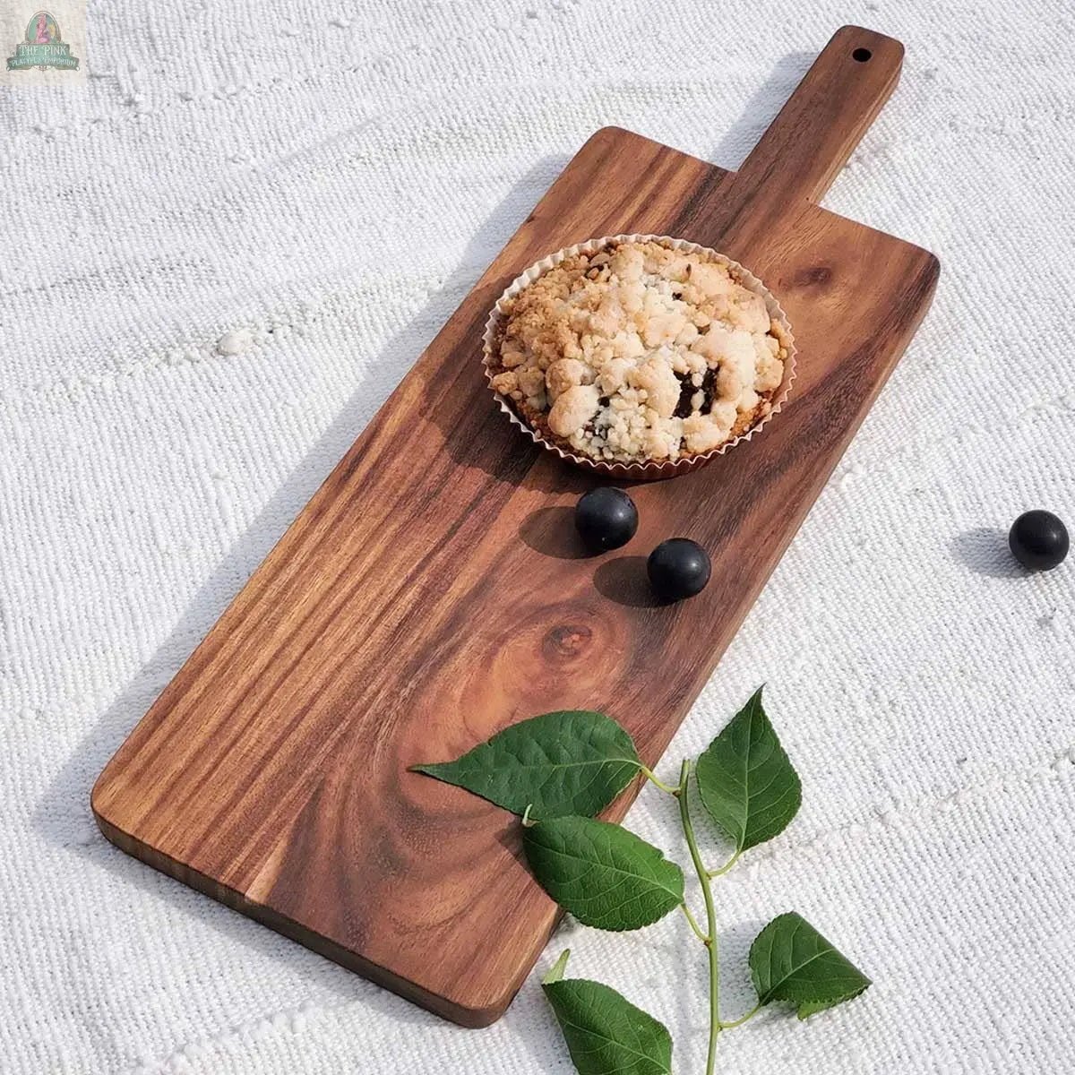 A crumb-topped muffin and two dark berries rest on the KORISSA Handmade Wooden Serving Board - Large, placed atop a textured white cloth with a green leafy stem nearby.