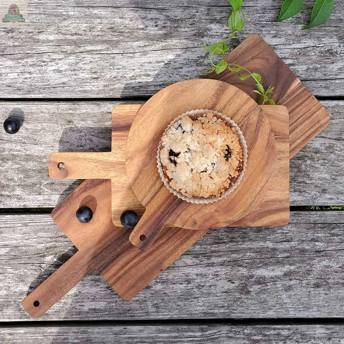 A blueberry muffin rests on the KORISSA Handmade Wooden Serving Board - Large, which is stacked over two rectangular boards on a rustic table, with three blueberries and a green vine nearby.