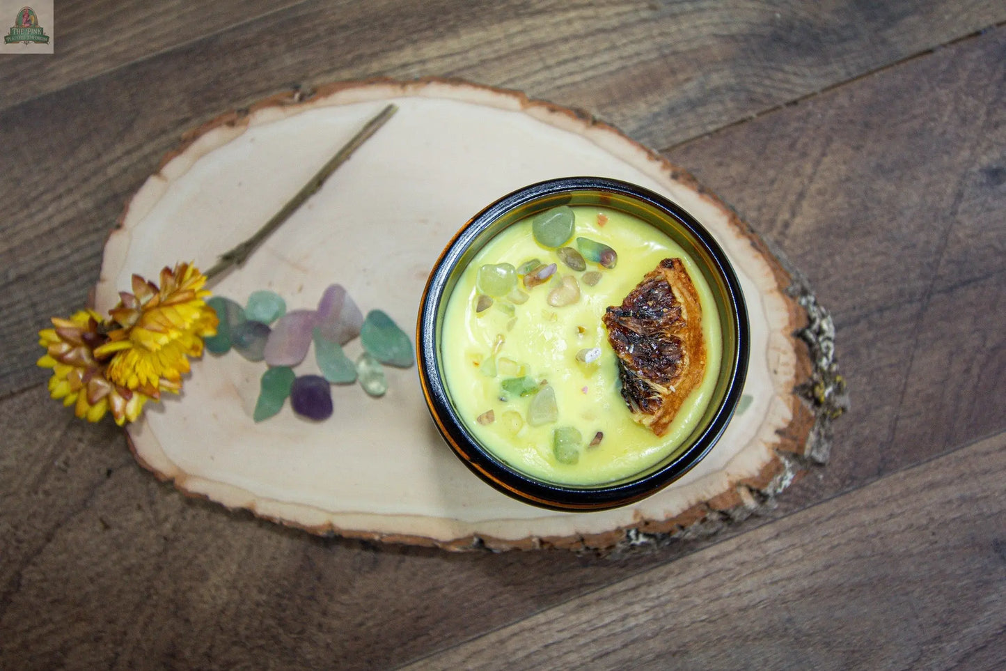 A bowl of yellow "Happiness" soup, garnished with charred orange and fresh herbs, sits on a wooden tray beside a yellow flower, colorful sea glass pieces, and a lemon verbena candle on a rustic wooden surface.