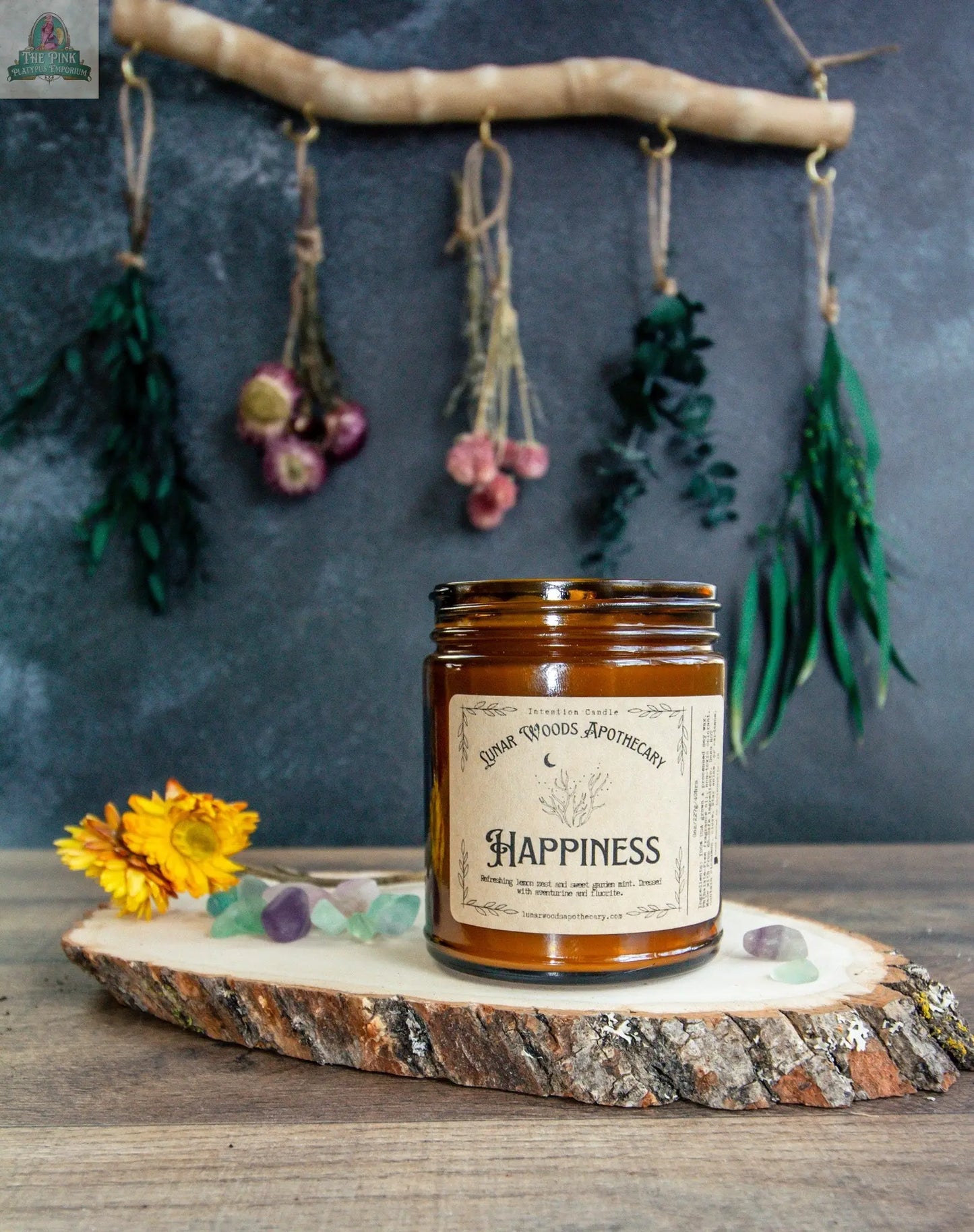 A brown glass jar labeled "Happiness" rests on a wooden slab, encircled by dried flowers, crystals, and hanging herbs. This candle creates a serene scene against a dark backdrop.