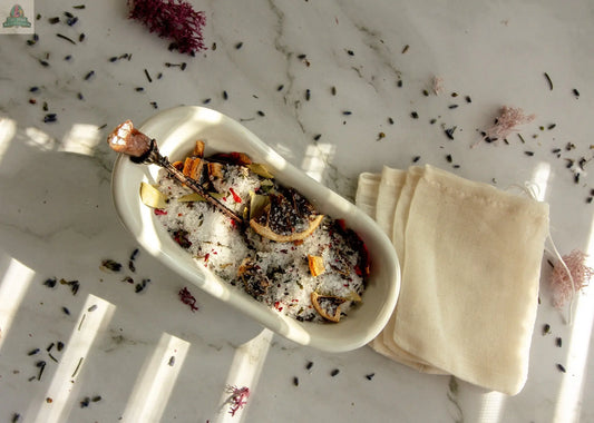 A white dish holds "Happiness," a blend of dried herbs, flowers, and salts for a soothing bath tea, set on marble with cream cloth bags. Sunlight creates striped shadows, highlighting this relaxing bath ritual kit.