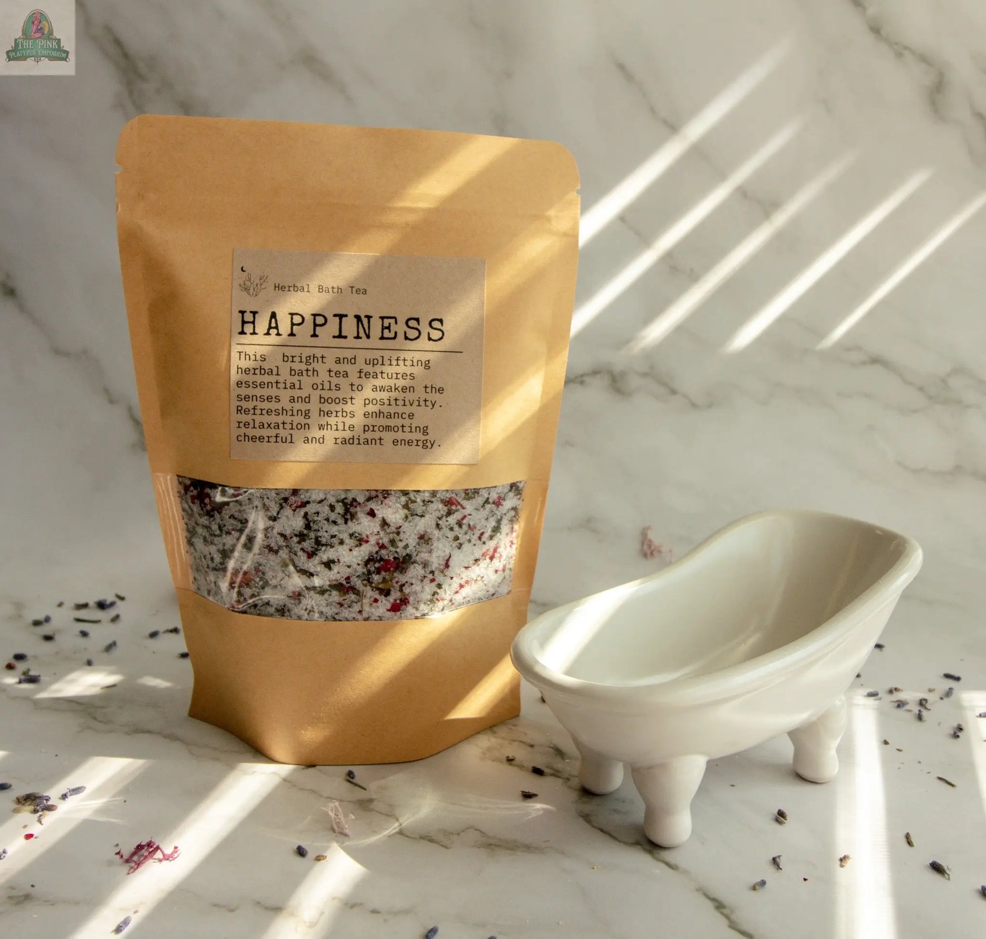 A brown paper bag labeled "Happiness," a herbal bath tea with orange and bergamot, stands beside a small white ceramic bathtub on a marble surface sprinkled with dried herbs, sunlight streaming in.