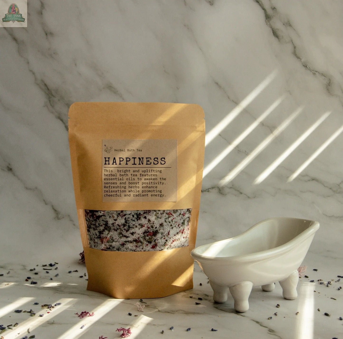 A brown paper pouch labeled Happiness, infused with orange and bergamot oils, stands beside a white clawfoot tub dish on marble, scattered dried herbs enhancing your self-care in diagonal sunlight.