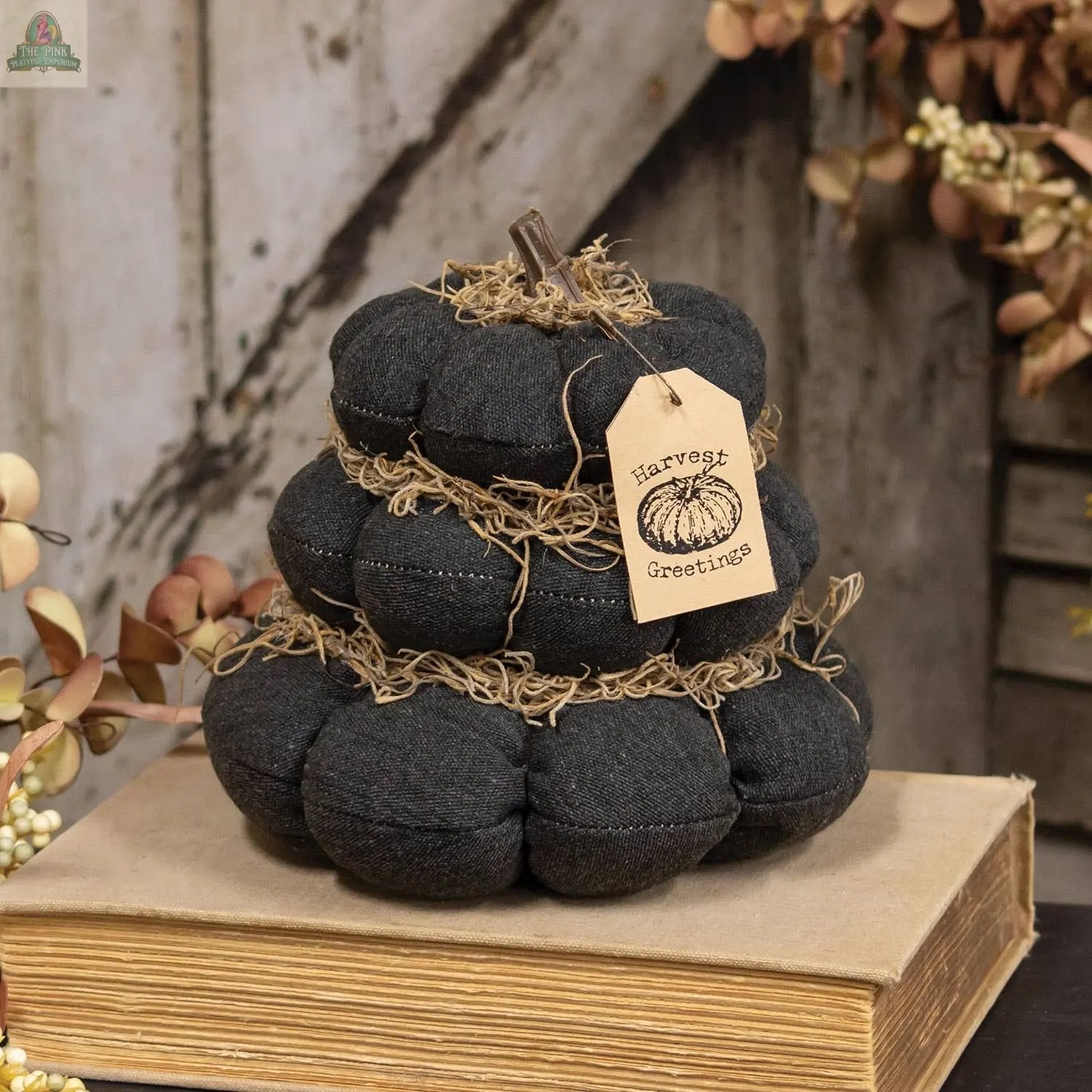 The Harvest Greetings Stuffed Pumpkin Stack features black plush pumpkins with a twig stem, straw accents, and a Harvest Greetings tag, resting on a closed book and surrounded by cozy autumn foliage for rustic decor.