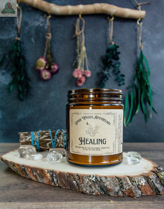 The Healing candle rests on a wooden slab amid crystals and sage bundles, with dried herbs in the background—creating a rustic, natural apothecary vibe that's perfect for energy clearing rituals.