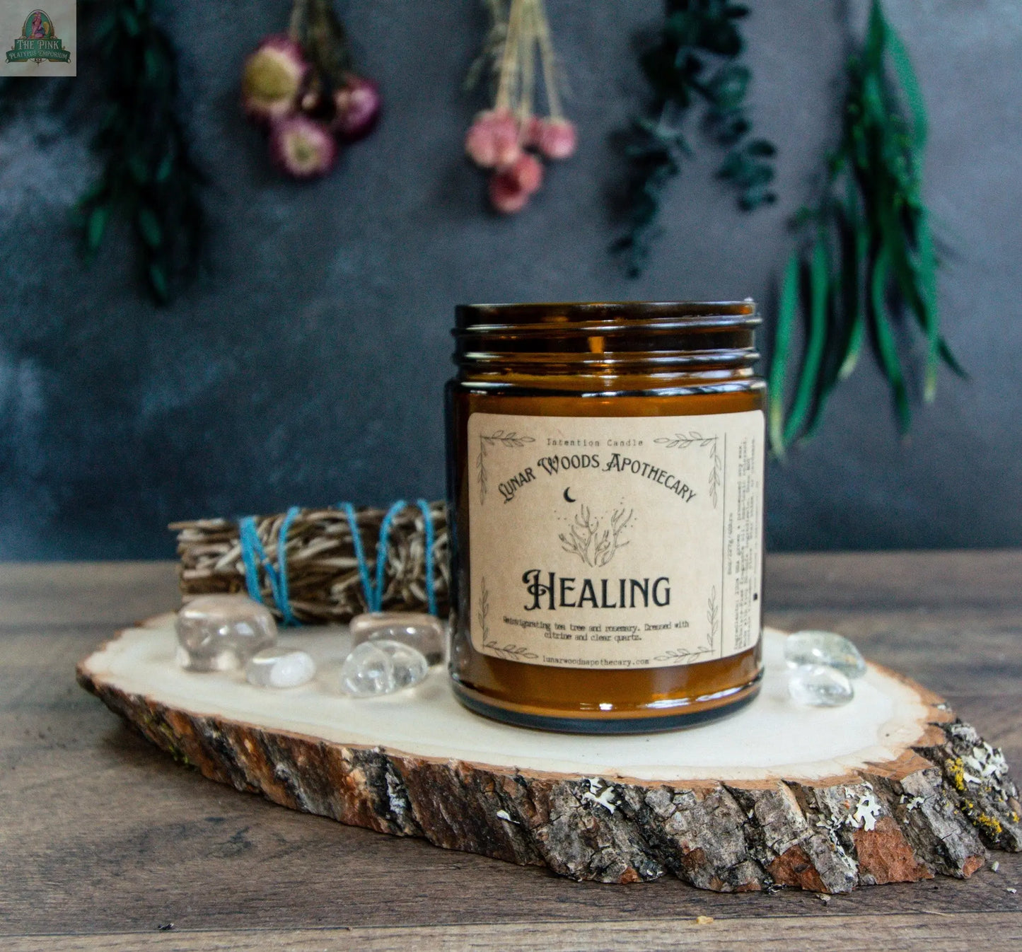 The Healing candle, in a brown glass jar, sits on a wood slice with clear stones and sage. Dried flowers and greenery hang above, creating a rustic, soothing energy cleansing display.