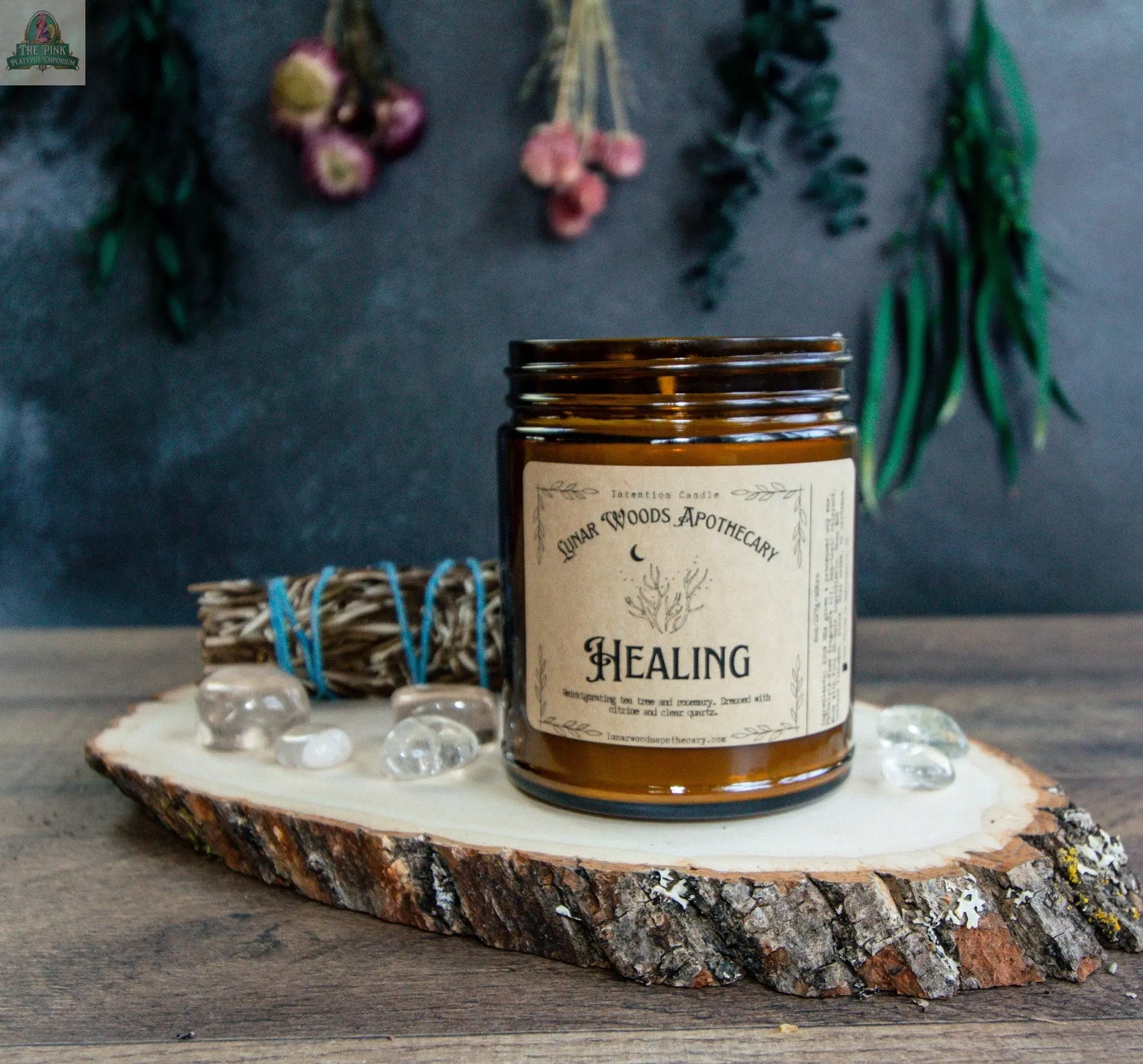 The Healing candle, in a brown glass jar, sits on a wood slice with clear stones and sage. Dried flowers and greenery hang above, creating a rustic, soothing energy cleansing display.