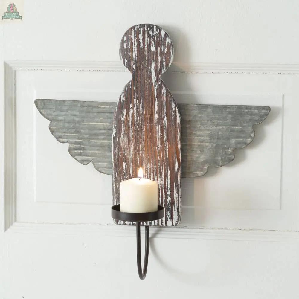 The Heavenly Angel Wall Sconce features a rustic distressed wood angel with corrugated metal wings, holding a lit white candle on a black metal candle holder, and is mounted on a white wall.