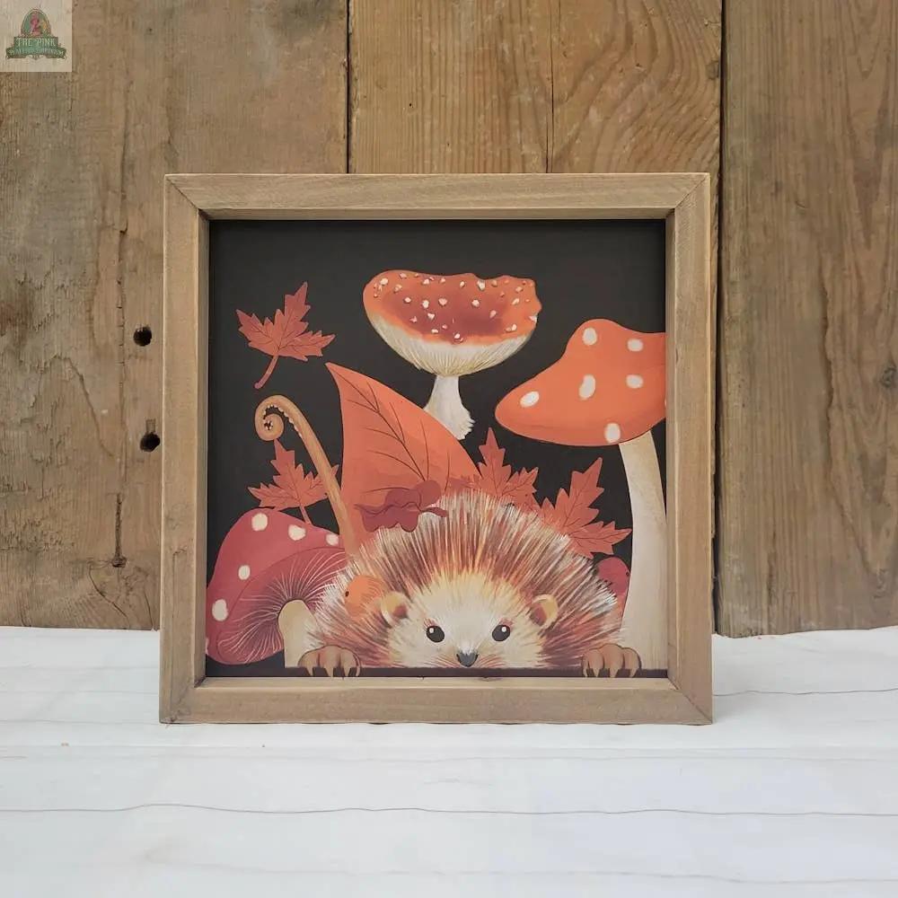 Hedgehog and Mushrooms in Frame features a cute hedgehog among mushrooms and autumn leaves on a dark background with a rustic wooden wall. Ideal as tabletop decor or wall art with its handy hanger.