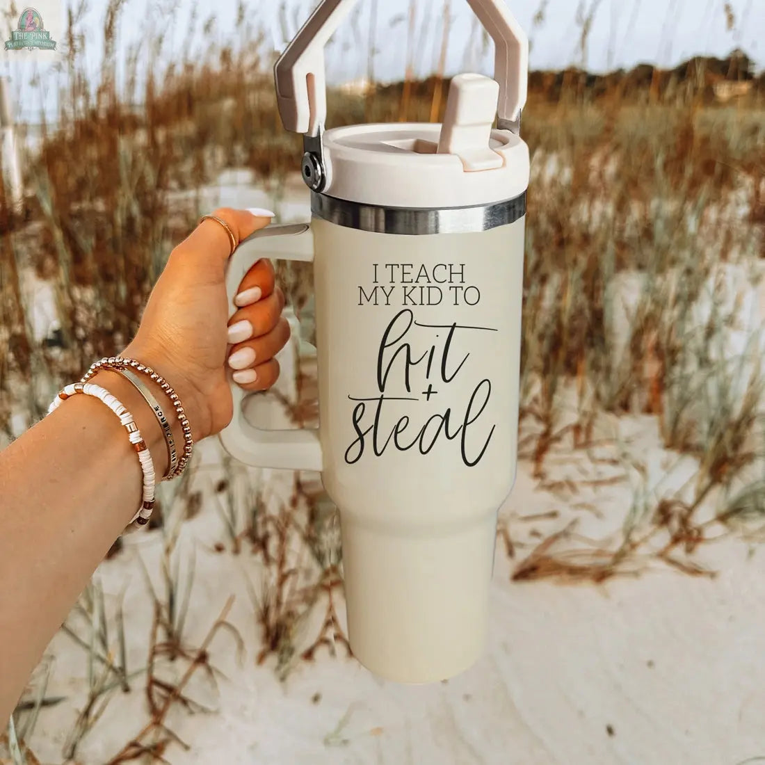 A hand holds the Hit & Steal cream-colored baseball mom tumbler with "I Teach My Kid to Hit + Steal" printed on it, set against a backdrop of beach and tall grass.