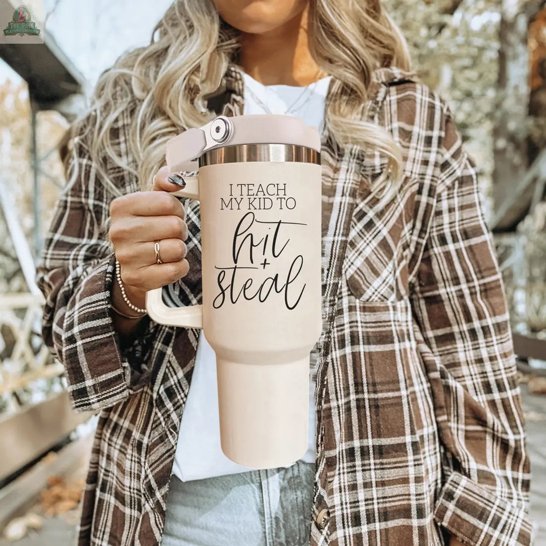 A woman in a brown plaid shirt holds a 40oz Hit & Steal tumbler, perfect for any baseball mom. Trees and a blurred outdoor background complete the scene.