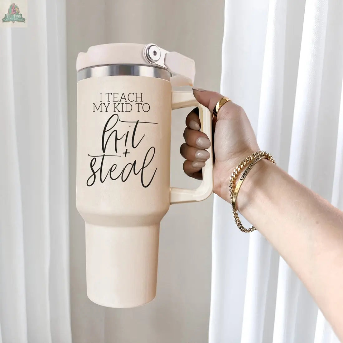 A hand holds the beige 40oz "Hit & Steal" tumbler with black script—ideal for any proud baseball mom’s collection. Gold rings, bracelets, and white curtains are in the background.