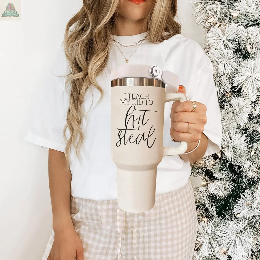 A woman stands by a snowy Christmas tree, holding the 40oz Hit & Steal baseball mom tumbler with “I Teach My Kid to Hit + Steal,” wearing a white shirt and checkered pants that show her love for baseball and her child.