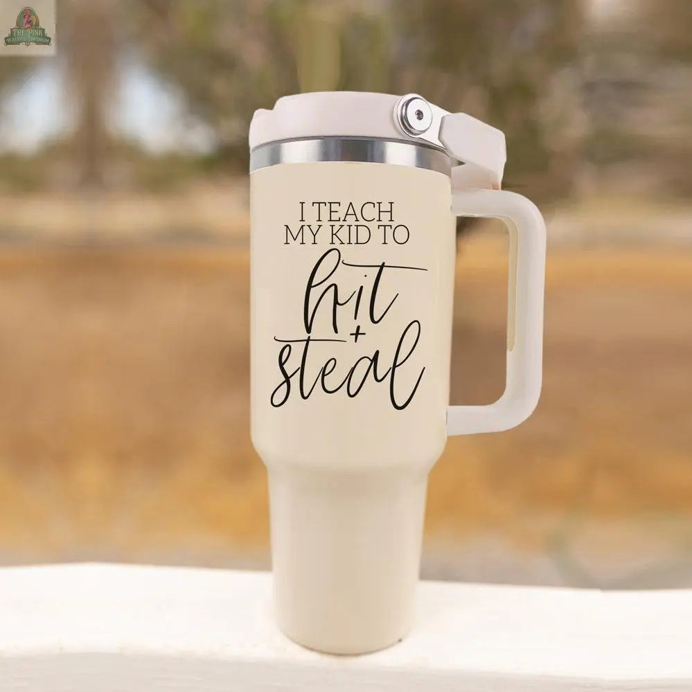 A 40oz beige Hit & Steal tumbler with a handle and silver lid sits on a white surface. "I TEACH MY KID TO hit + steal" is printed in black, making it the perfect baseball mom mug for outdoor adventures. The background is blurred outdoors.