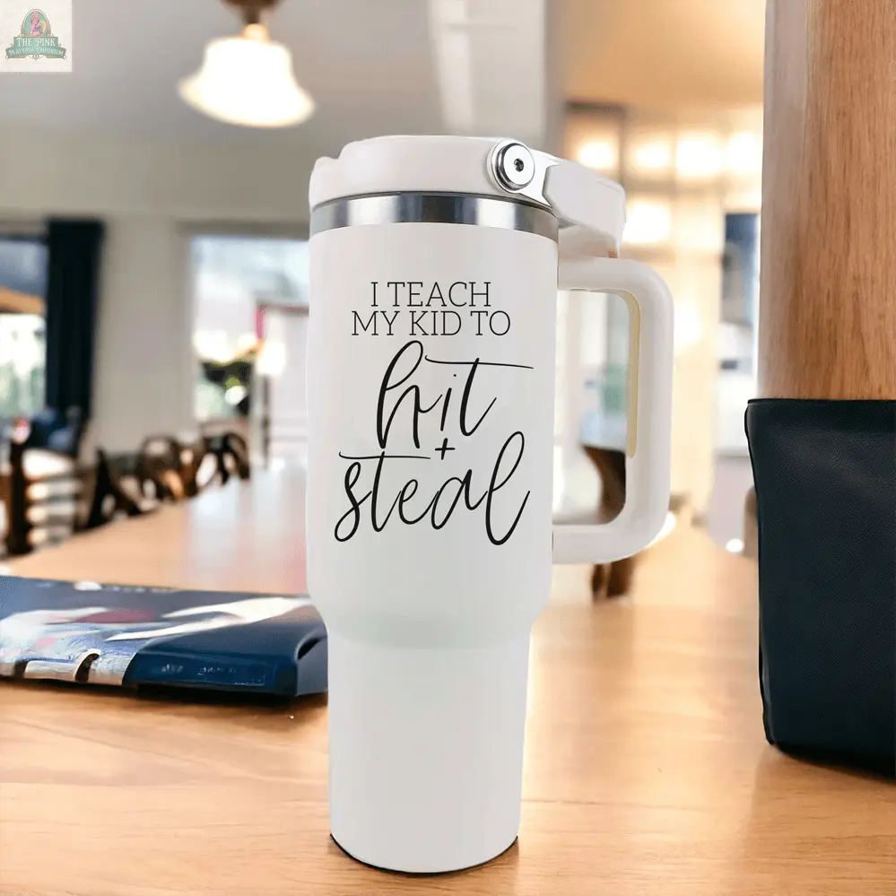 A white “Hit & Steal” baseball mom tumbler sits on a wooden table, with “I Teach My Kid to Hit + Steal” in black script. The cozy dining room background highlights this perfect 40oz tumbler for sports-loving moms.