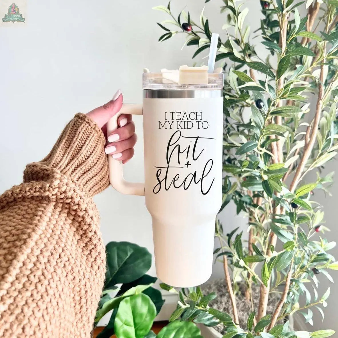 A person in a brown chunky sweater holds a 40oz Hit & Steal tumbler with "I Teach My Kid to Hit + Steal" printed on it, set against green leafy plants.