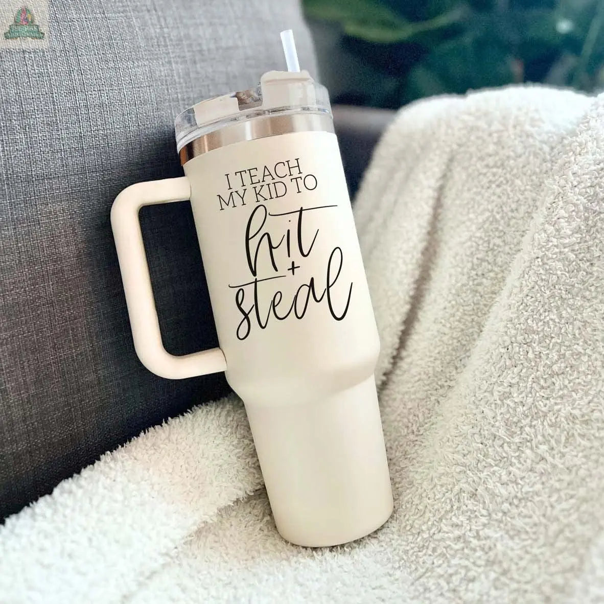 A white 40oz Hit & Steal baseball mom tumbler with a handle and straw sits on a blanket. The tumbler features “I Teach My Kid to Hit + Steal” in decorative black lettering.