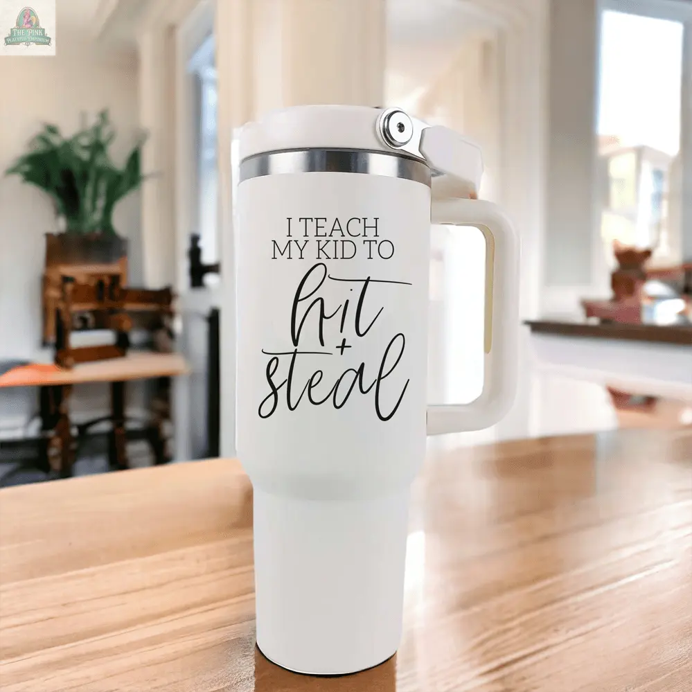 The "Hit & Steal" 40oz white tumbler with handle rests on a wooden table, featuring the phrase "I Teach My Kid to Hit + Steal"—an ideal baseball mom mug for sunny, plant-filled rooms.
