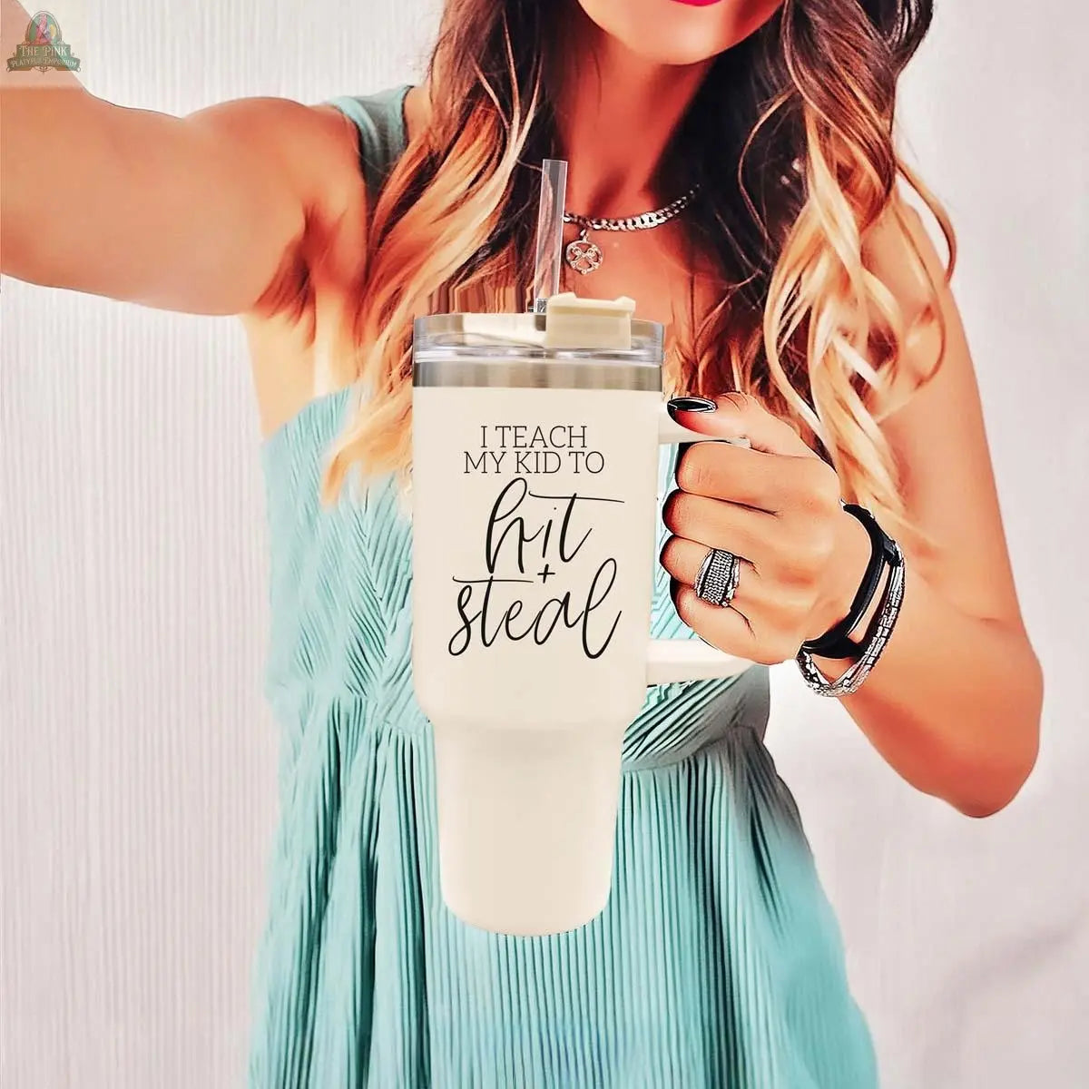 A woman in a blue sleeveless dress holds the 40oz Hit & Steal baseball mom tumbler with "I Teach My Kid to Hit + Steal" text, sporting a ring and smartwatch against a light, plain background.
