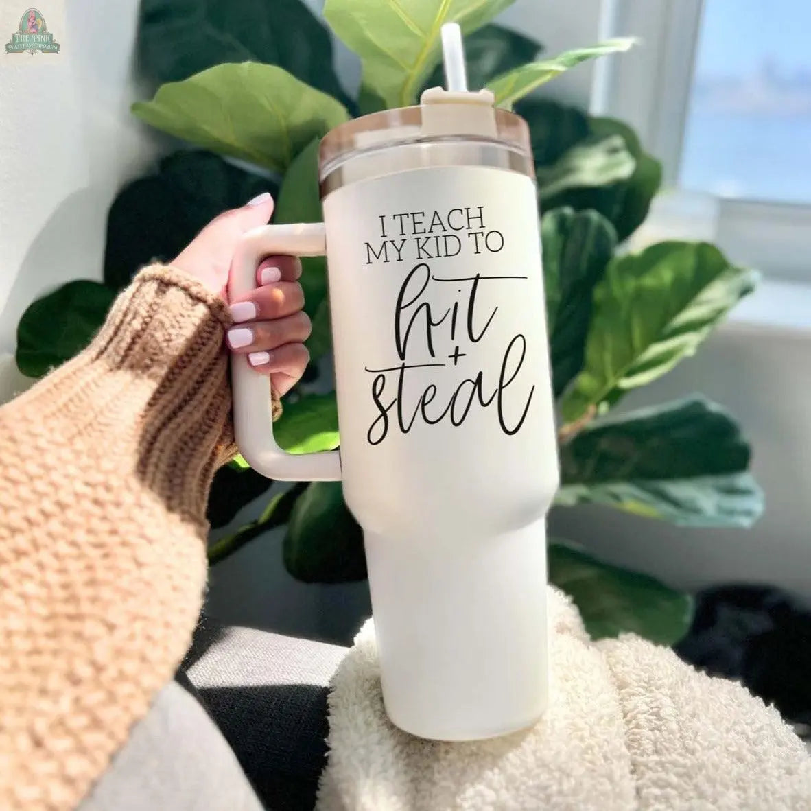 A person in a tan knit sweater holds a 40oz "Hit & Steal" baseball mom tumbler with a straw. Sunlight from a nearby window shines on a leafy green plant.