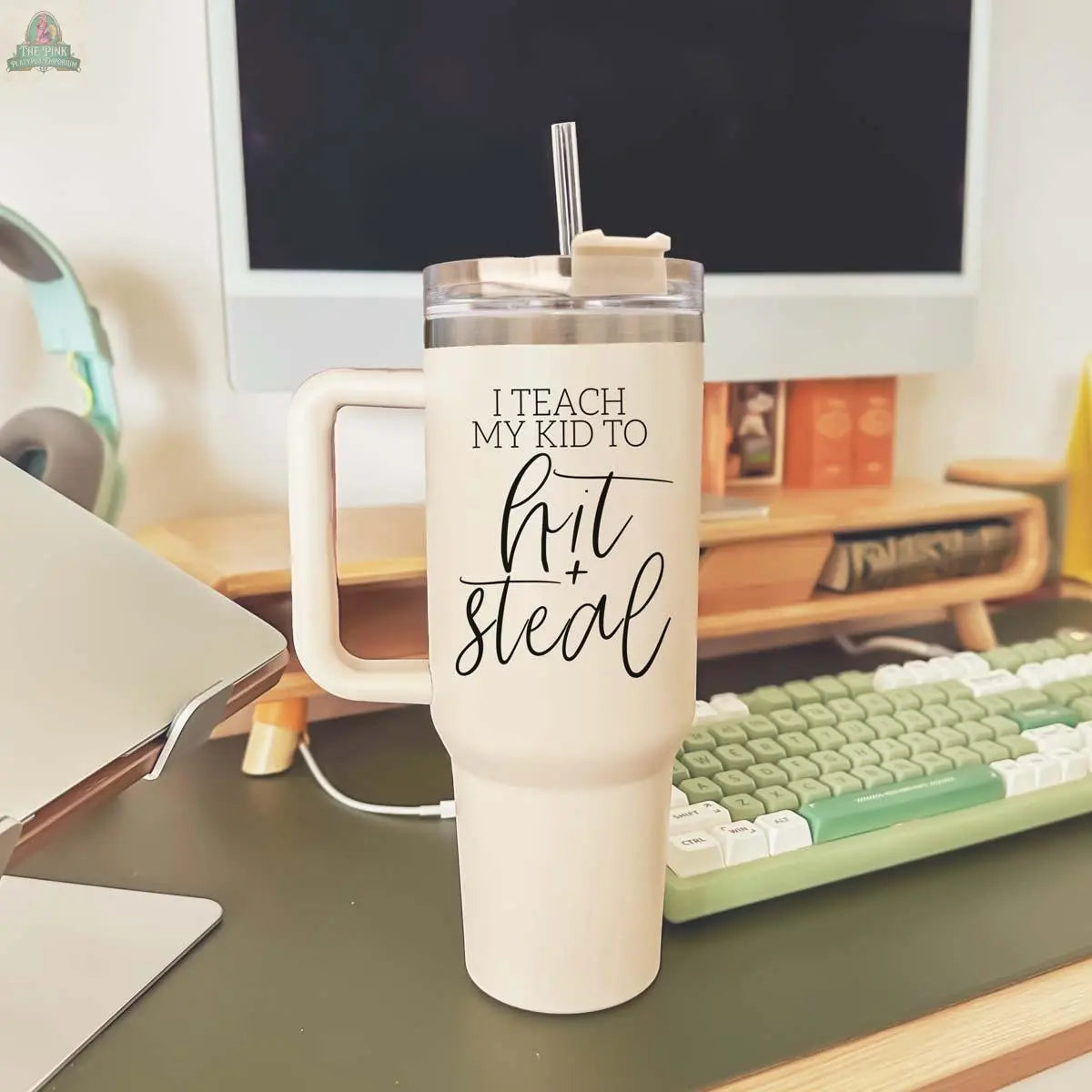 A beige 40oz Hit & Steal tumbler with a straw, featuring "I Teach My Kid to Hit + Steal" in black script, sits on a desk next to a green-and-white keyboard, laptop, headphones, and monitor.