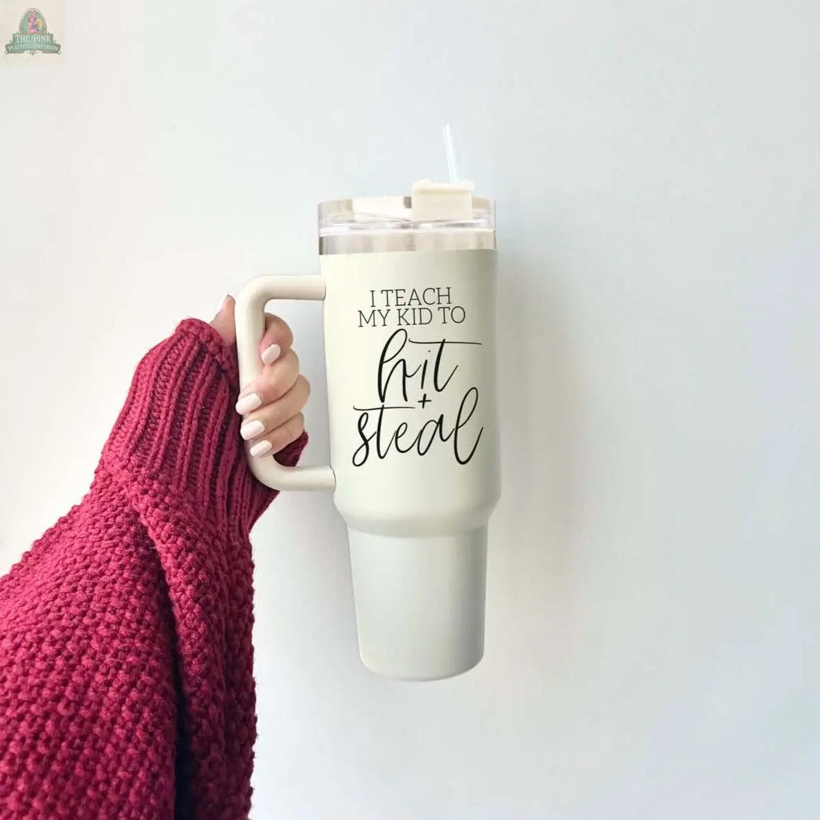 A hand in a red knit sweater holds the Hit & Steal 40oz baseball mom tumbler with “I Teach My Kid to Hit + Steal” in black script. It has a clear lid, a straw, and a plain white background.